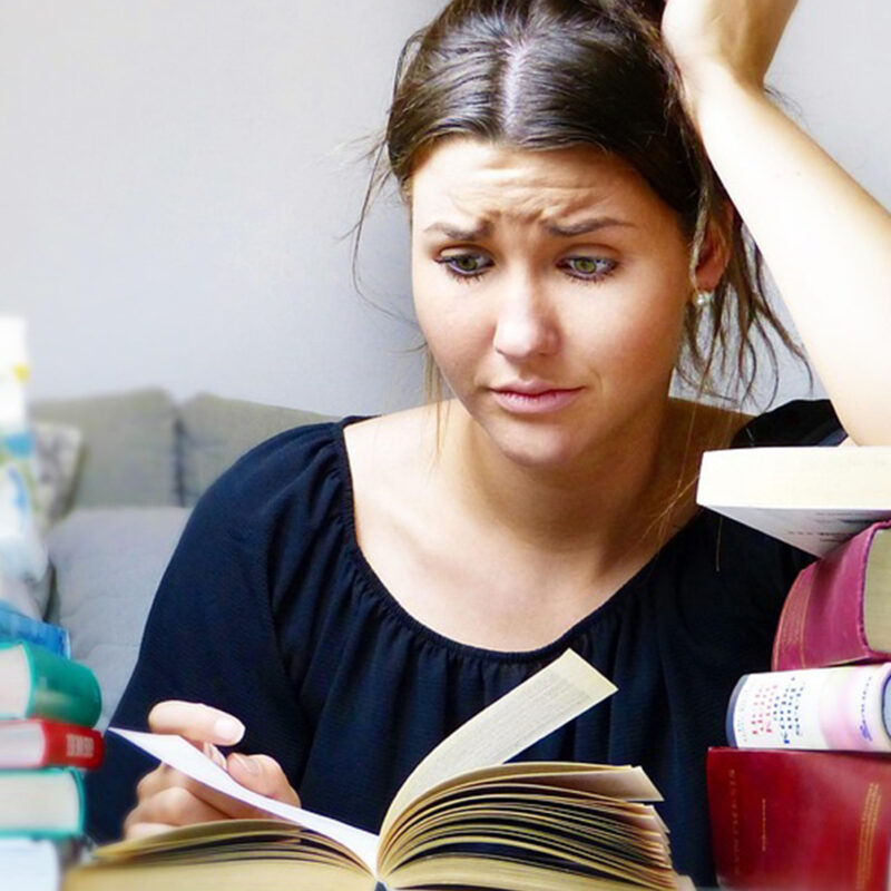 Confused Female Student Reading with Stacks of Textbooks